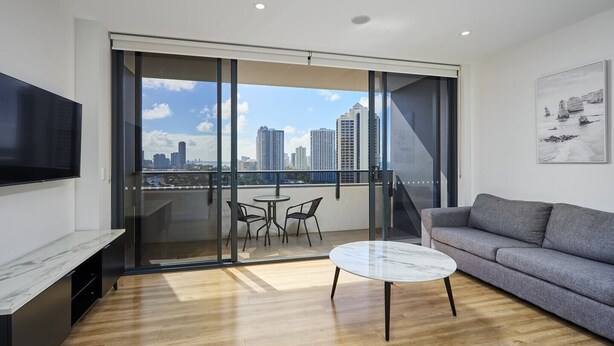 Un salon moderne avec un canapé gris, une table basse ronde et un téléviseur fixé au mur. Il y a un balcon avec une porte vitrée donnant sur un espace extérieur offrant une vue sur la ville.