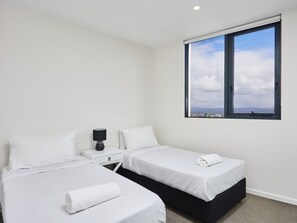 A hotel room with two single beds, a nightstand with a lamp, and a large window offering a city view. - Partially Oceanview 2 bedroom apartment in Surfers (Surfers Paradise)