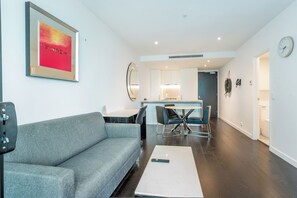 A modern living room with a grey sofa, a dining table with four chairs, a kitchen area with a fridge, and a clock on the wall.