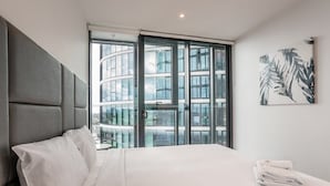 Moderni hotellihuone, jossa on suuri sänky, pääty, näkymä lasiseen rakennukseen ja seinälle asennettu taideteos.
