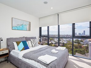 A modern bedroom with a large bed, a city view through floor-to-ceiling windows, and a painting on the wall. - Comfy 2BR Apartment Central Surfers w/Parking (Surfers Paradise)