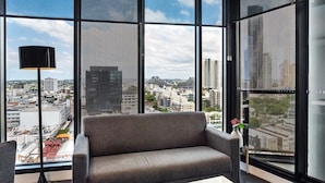 A modern living room with a grey sofa, a small coffee table, and a floor lamp. Large windows offer a city view. - City View 1BR Apartment + ParkingandWalk to Station (Fortitude Valley)