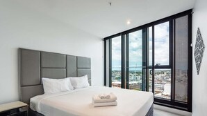 A modern hotel room with a large bed, a nightstand, and a view of the city through large windows. - City View 1BR Apartment + ParkingandWalk to Station (Fortitude Valley)