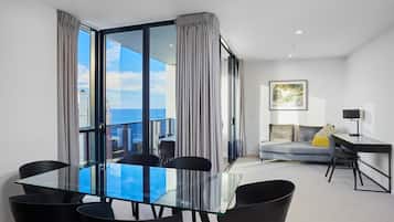 A modern hotel room with a glass dining table, black chairs, a grey sofa, and a city view.