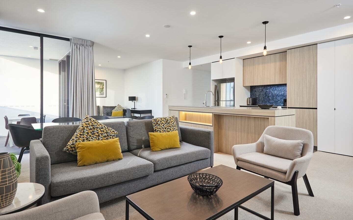 A modern living room with a grey sofa, yellow cushions, a coffee table, and a dining area with a kitchen in the background.