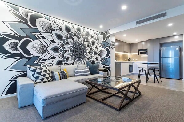A modern living room with a large abstract wall art, a sofa, a coffee table, and a kitchen area in the background.