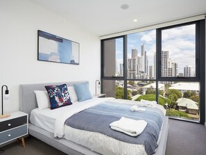A modern bedroom with a large bed, a nightstand, a bedside table, and a view of a cityscape through large windows. - Iconic Surfer's Sweet one bedroom Home in Surfers (Surfers Paradise)