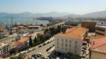 Un paysage urbain côtier avec un imposant bâtiment blanc, un port animé avec des grues et des navires, et une vue dégagée sur la mer et les montagnes en arrière-plan.
