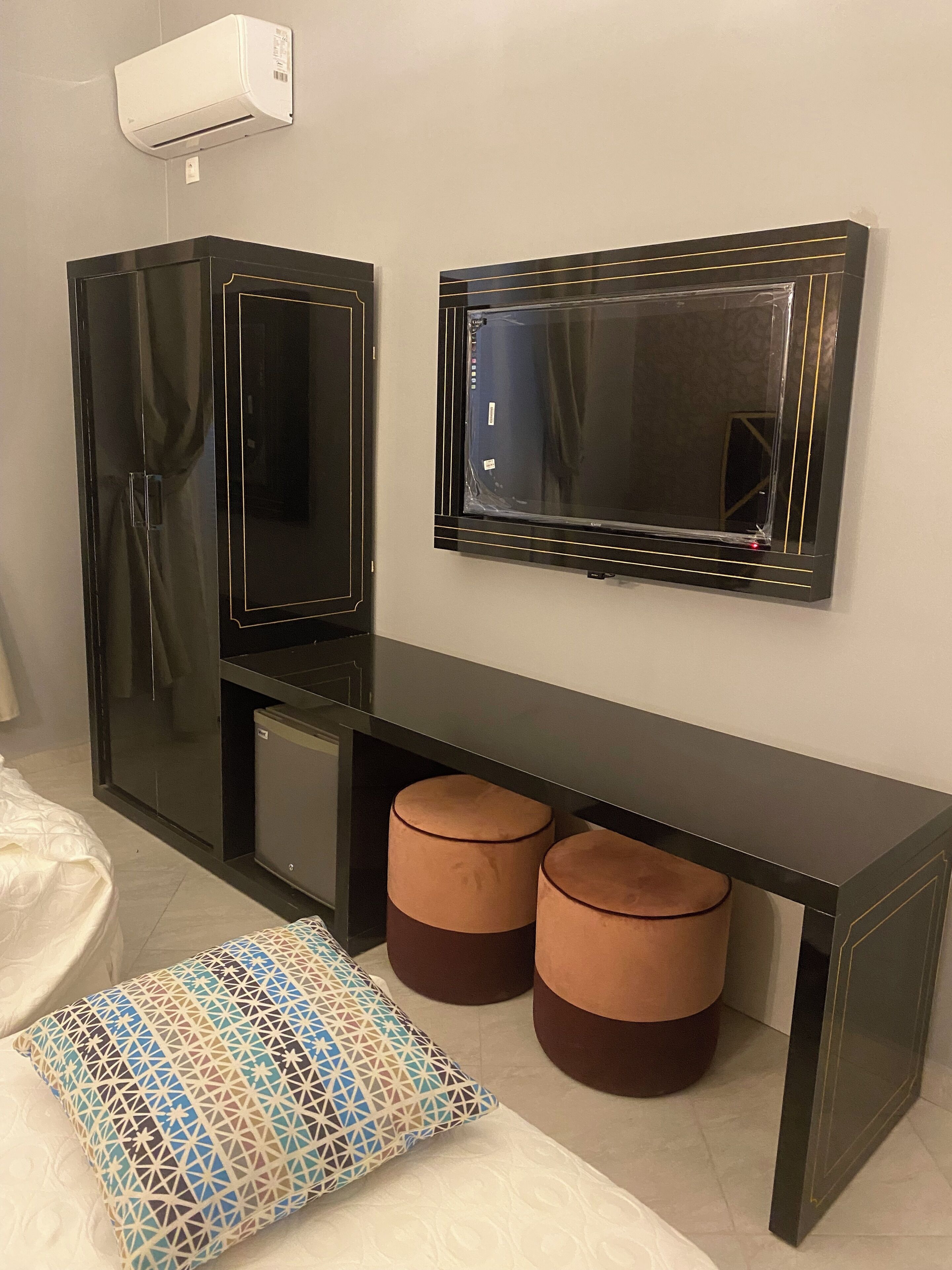 A modern room with a black console table, two cushioned stools, a patterned pillow, a large mirror, and a wall-mounted air conditioner.