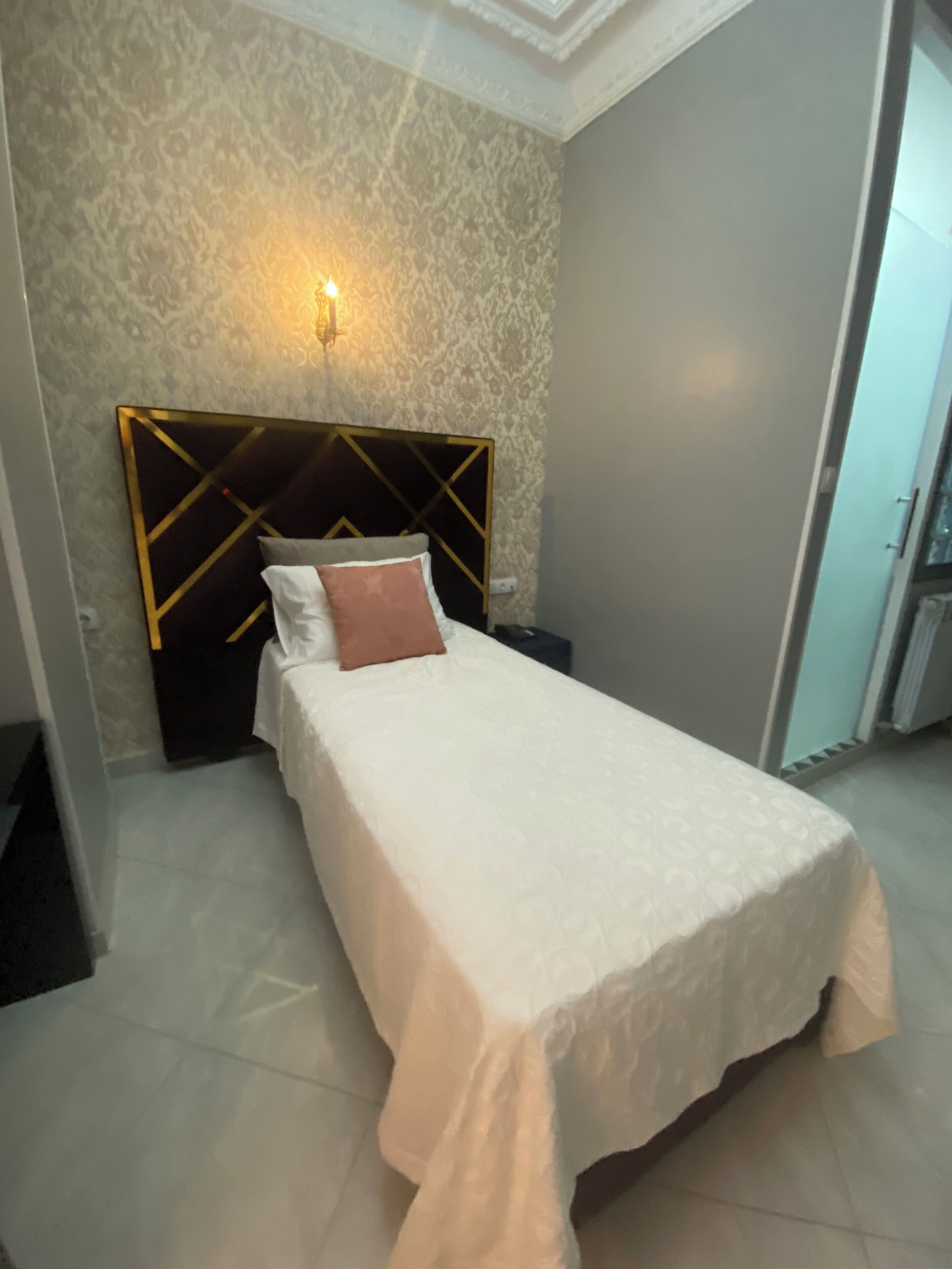 A neatly made bed with a patterned headboard, a wall-mounted light fixture, and a tiled floor.