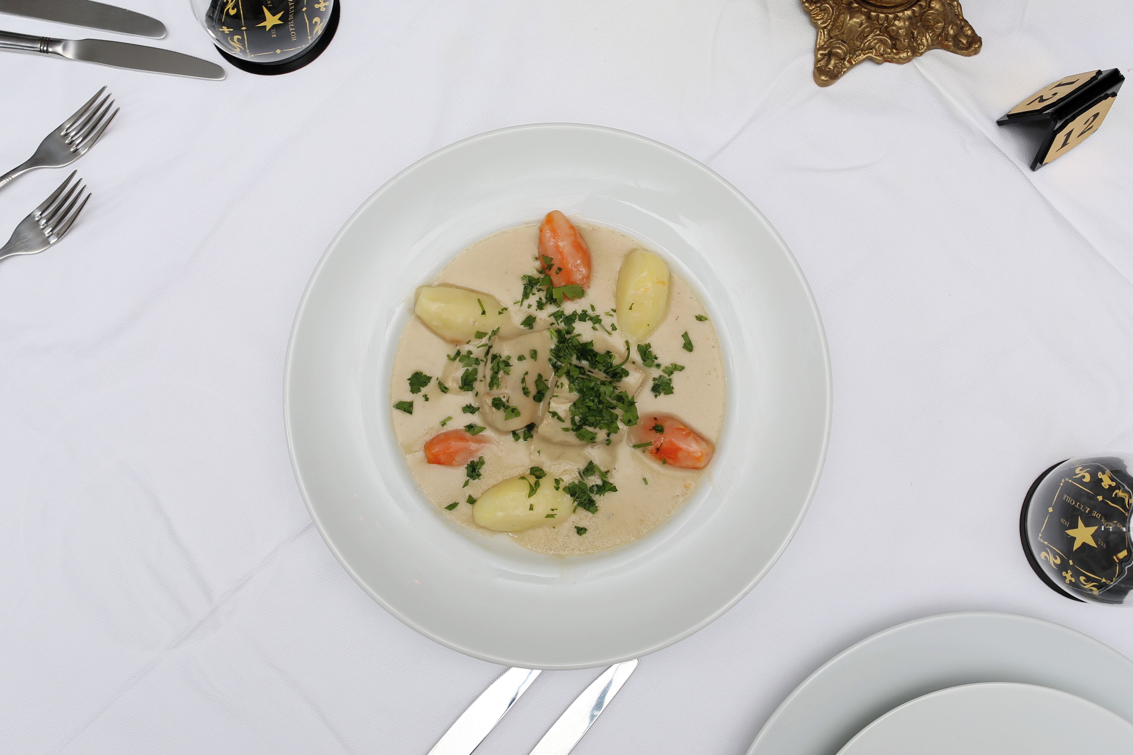 Une table dressée avec une nappe blanche, une assiette de soupe garnie d’herbes aromatiques, ainsi que des couverts et un objet décoratif.