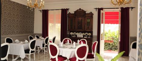 Un comedor con mesas y sillas blancas, una araña y vistas al exterior.