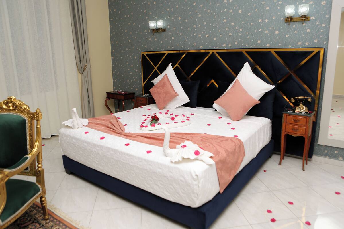 A neatly made bed with rose petals and a swan decoration, a gold-framed chair, and a classic wall clock.