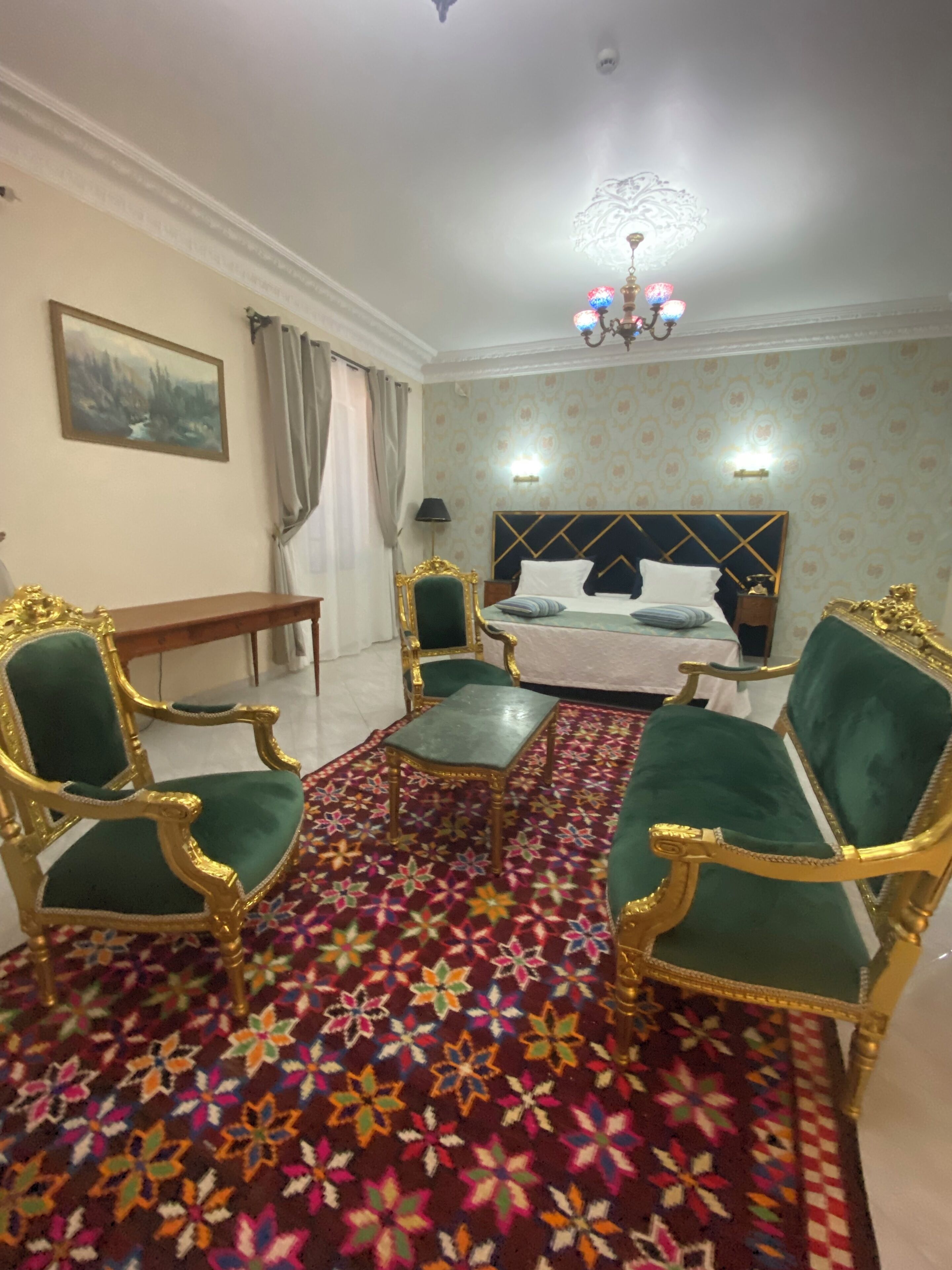 Una habitación con una cama, dos sillones verdes tapizados, una mesita, una alfombra estampada, un cuadro en la pared y una araña.