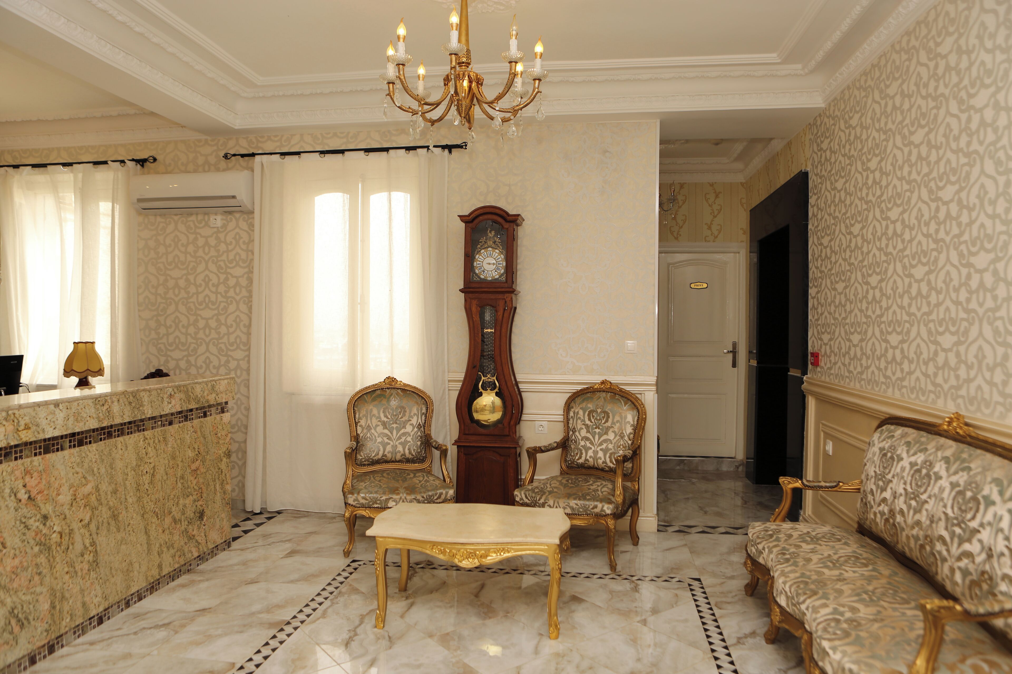 A formal sitting room with a marble floor, a chandelier, two ornate chairs, a table, and a grandfather clock.