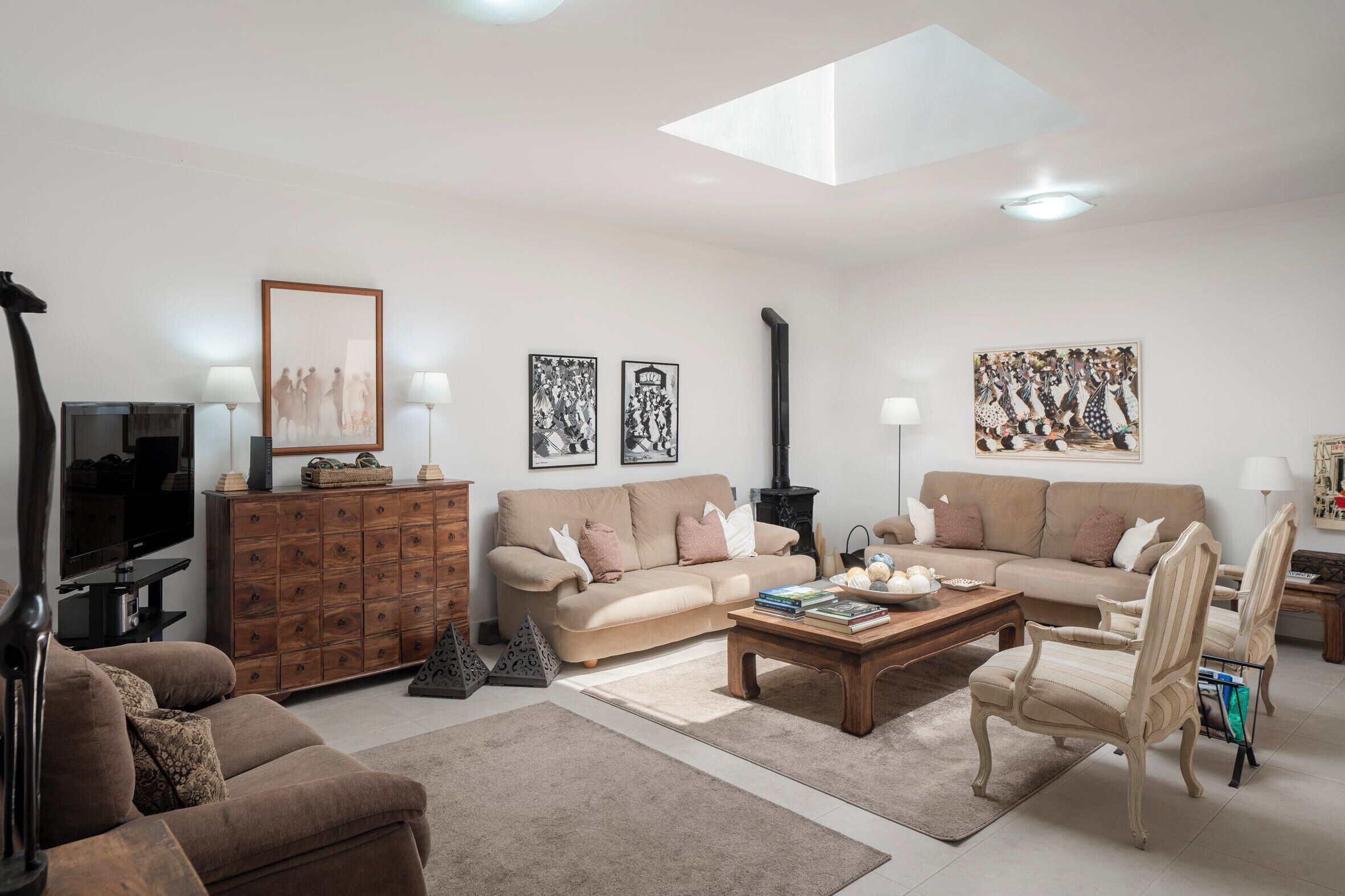 A living room with a sofa, armchairs, a coffee table, and a TV. There are wall-mounted lamps, a fireplace, and framed pictures.