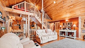 A spacious living area with a high ceiling, wooden walls, a sofa, a dining table, and a staircase leading to an upper level.