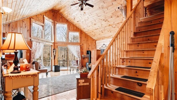 A wooden cabin interior with a staircase, large windows, and a ceiling fan.