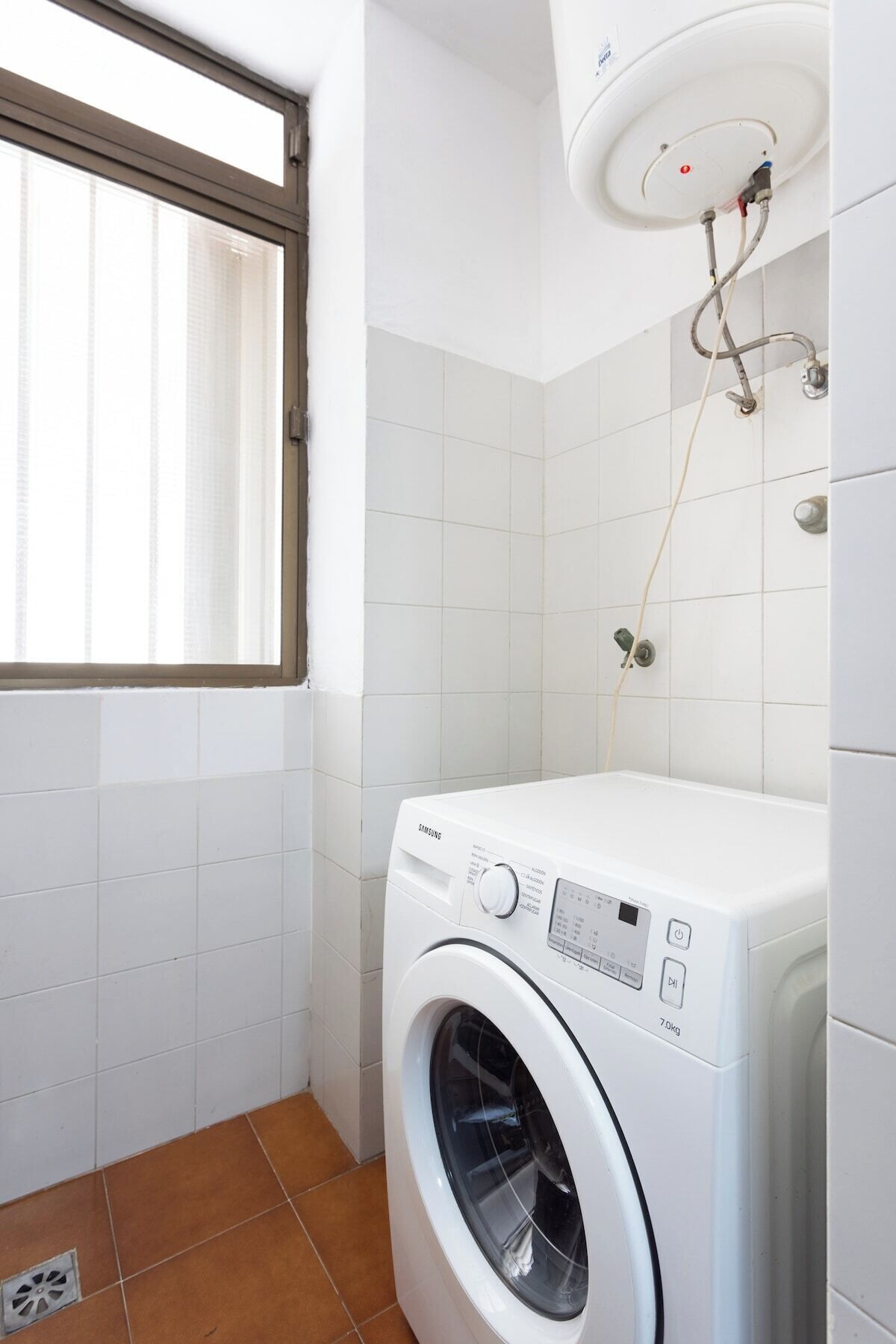Een kleine wasruimte met een voorlader wasmachine, een raam met zonwering en een aan het plafond bevestigde boiler.