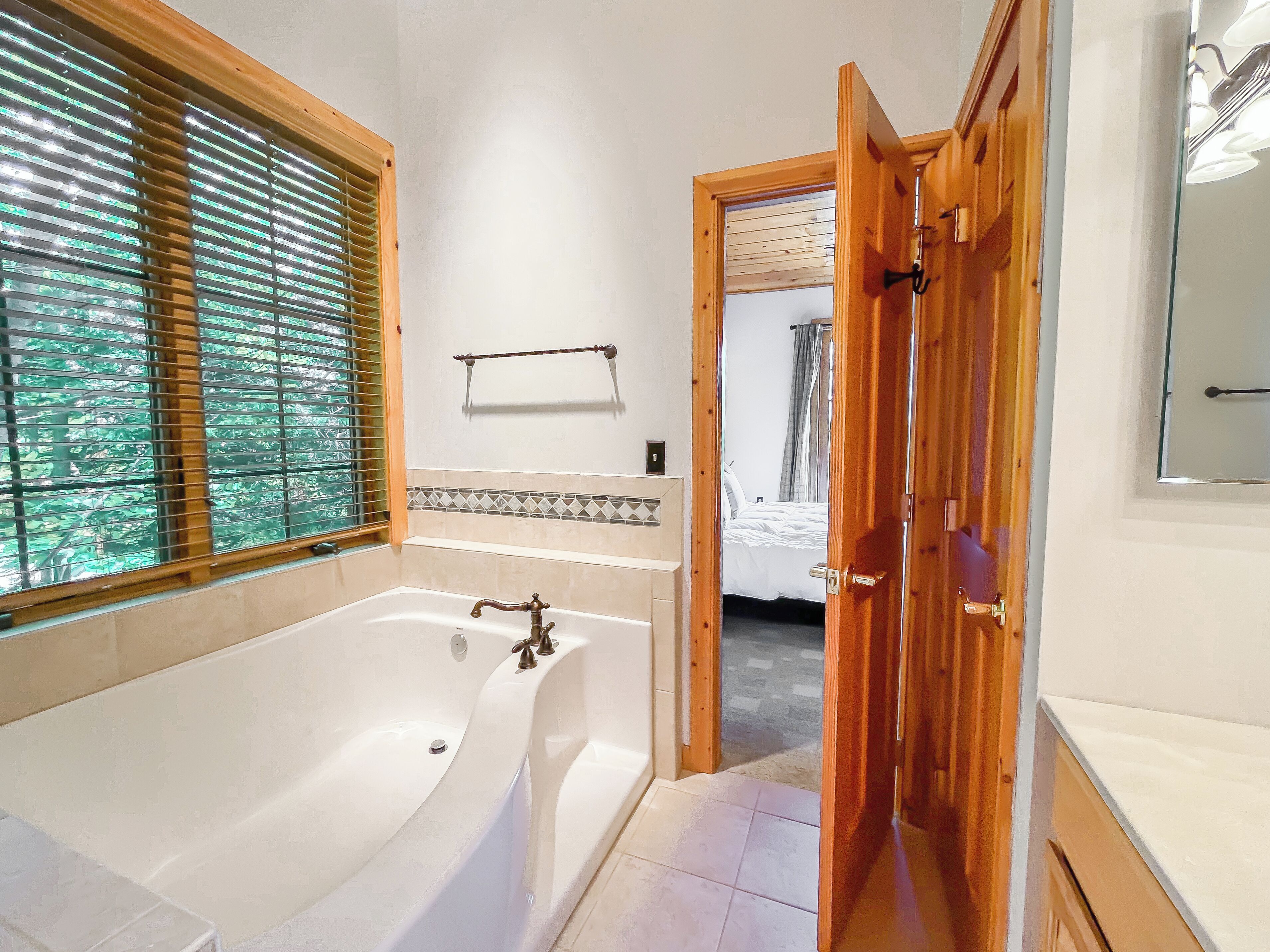 Un baño con una bañera grande, persianas de madera, un perchero y una habitación visible a través de una puerta abierta.