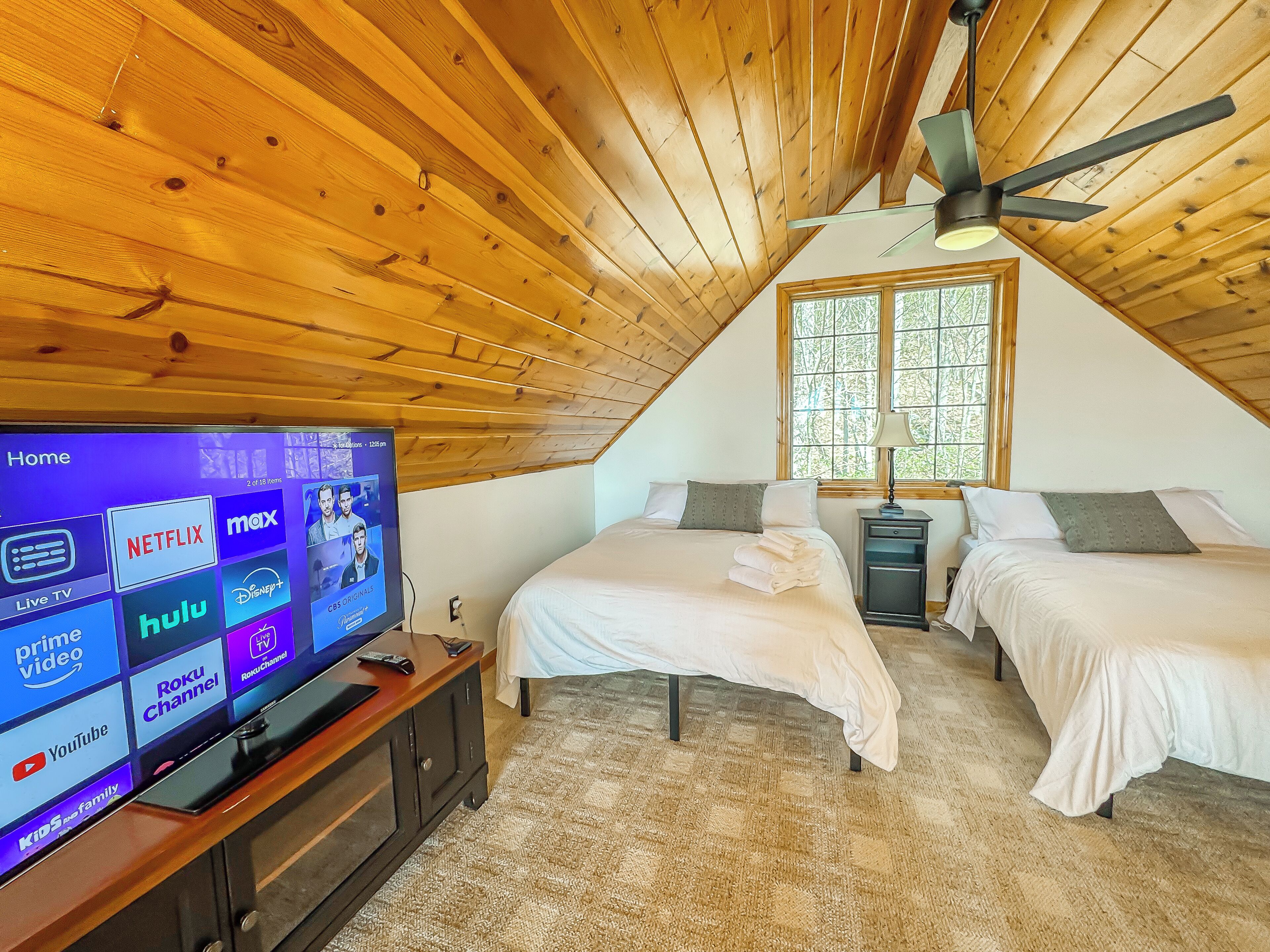 A bedroom with a TV displaying various streaming services, two beds, a nightstand, a lamp, and a ceiling fan.