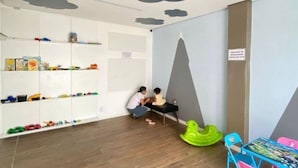 A room with a wooden floor, a white shelving unit with toys, a green and pink chair, and a table with a colorful tablecloth. - Apartamento Park Quinet  (Juiz de Fora)
