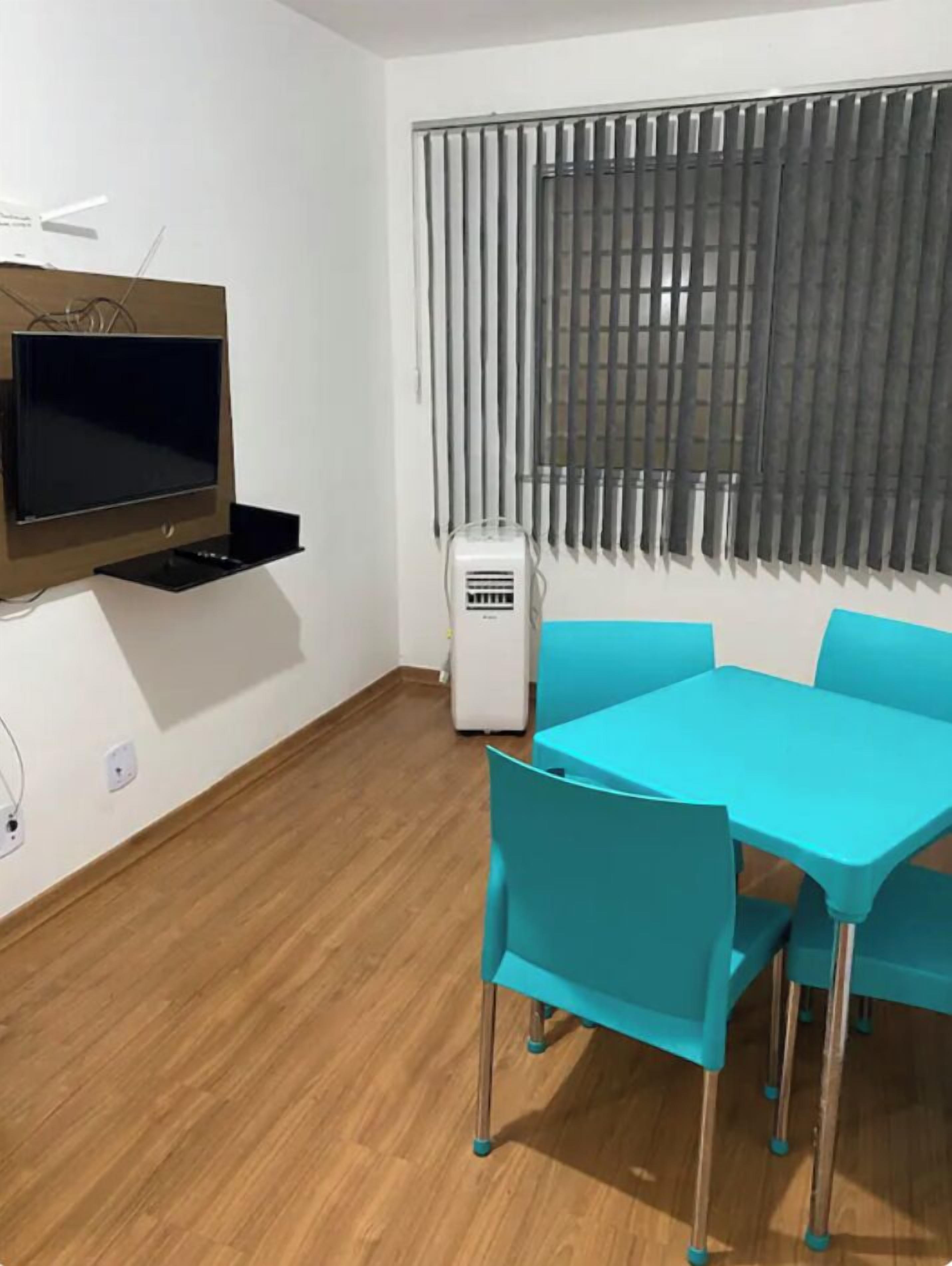 Quarto com piso de madeira, televisão na parede, ar-condicionado branco e mesa azul com quatro cadeiras.