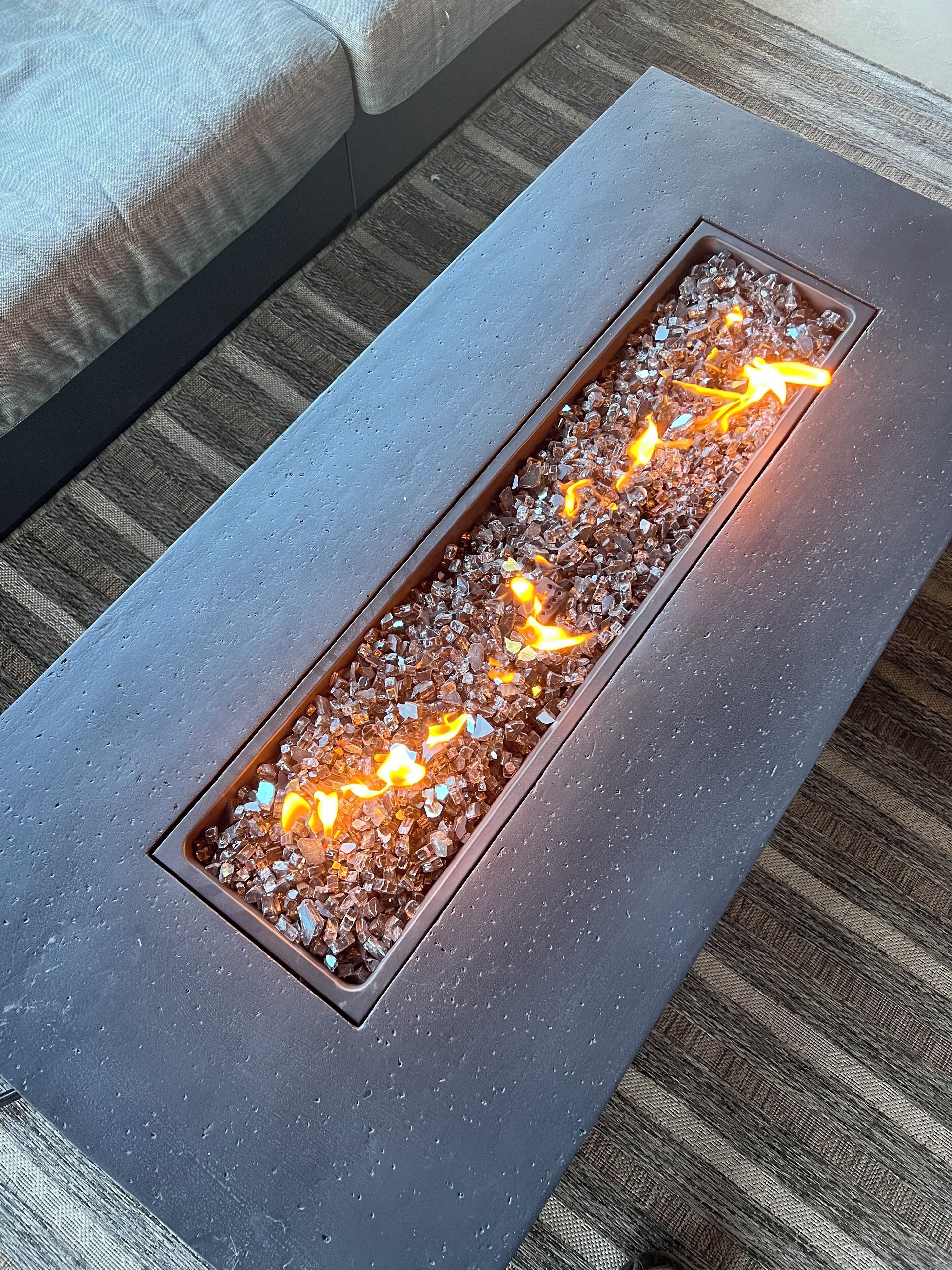 A rectangular fire feature with a metal frame and glass sides, placed on a textured grey surface.