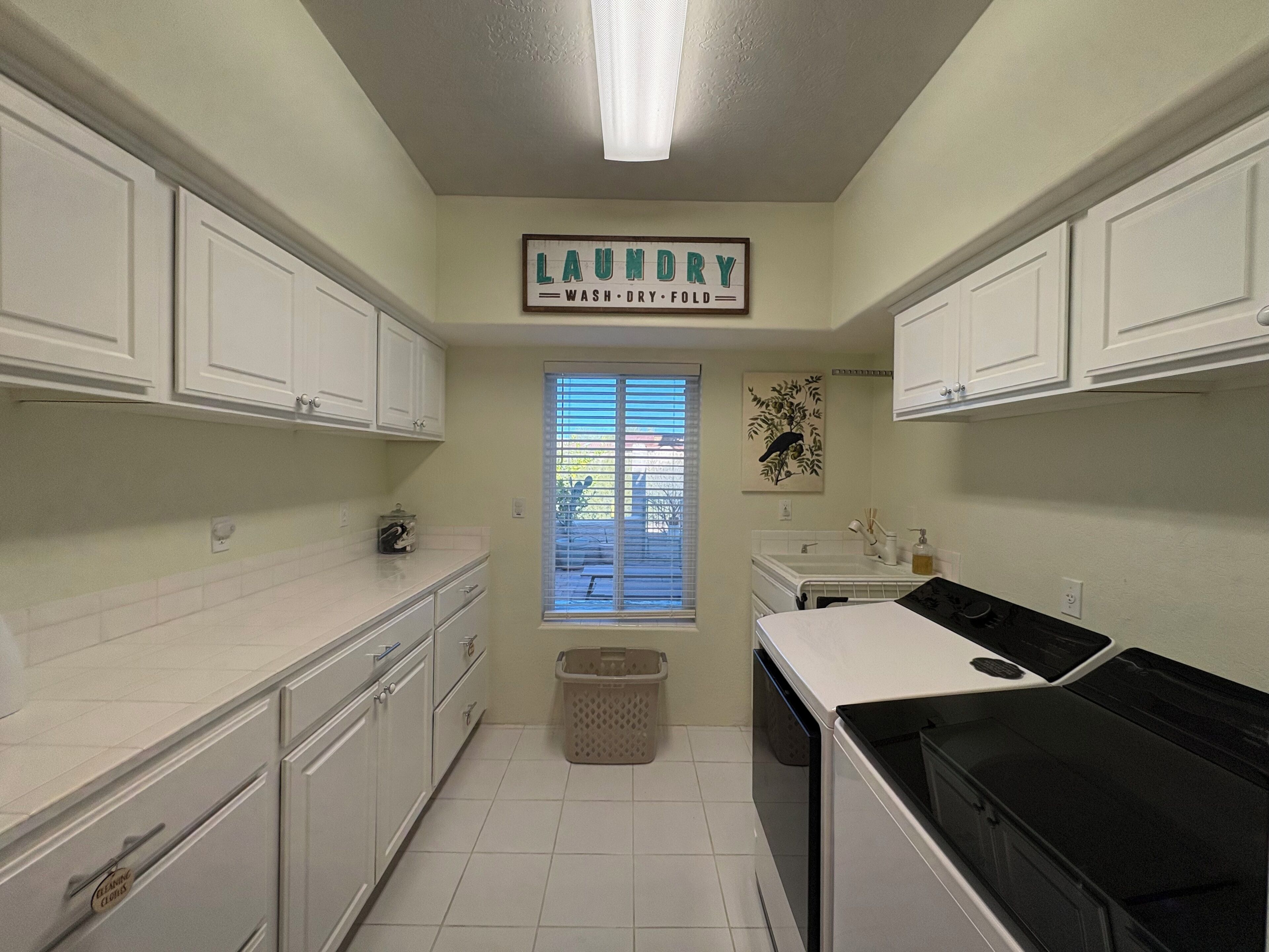 A laundry room with white cabinets, a washing machine, and a dryer. There is a window with blinds and a picture on the wall.