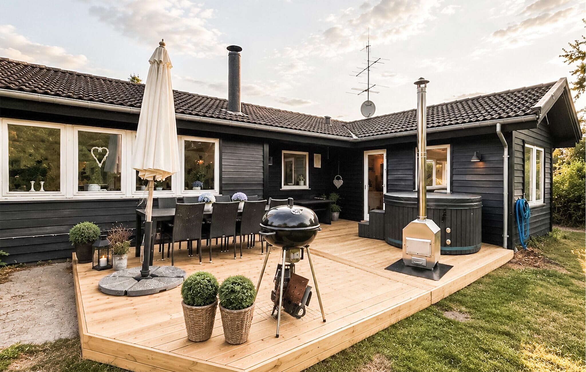 Une maison noire avec une terrasse en bois, un barbecue, un jacuzzi et du mobilier d’extérieur.