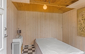 Quarto pequeno com uma cama de solteiro, paredes de painel de madeira, radiador, luminária de mesa e chão xadrez.