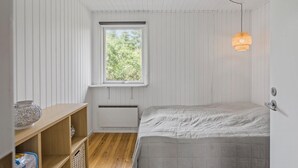 A small, bright room with a single bed, a window with a view of greenery, a shelf with a decorative item, and a hanging light fixture.