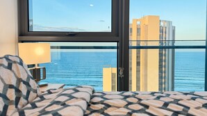 Habitación de hotel con cama, mesita de noche, lámpara y un amplio ventanal que ofrece vistas al mar y edificios.