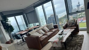 Una sala de estar moderna con un sofá de cuero marrón, una mesa de comedor y vistas a la ciudad y al océano a través de amplios ventanales.