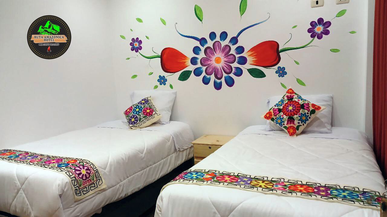A hotel room with two beds, each with a colorful embroidered bedspread. There is a wall mural with flowers and leaves.