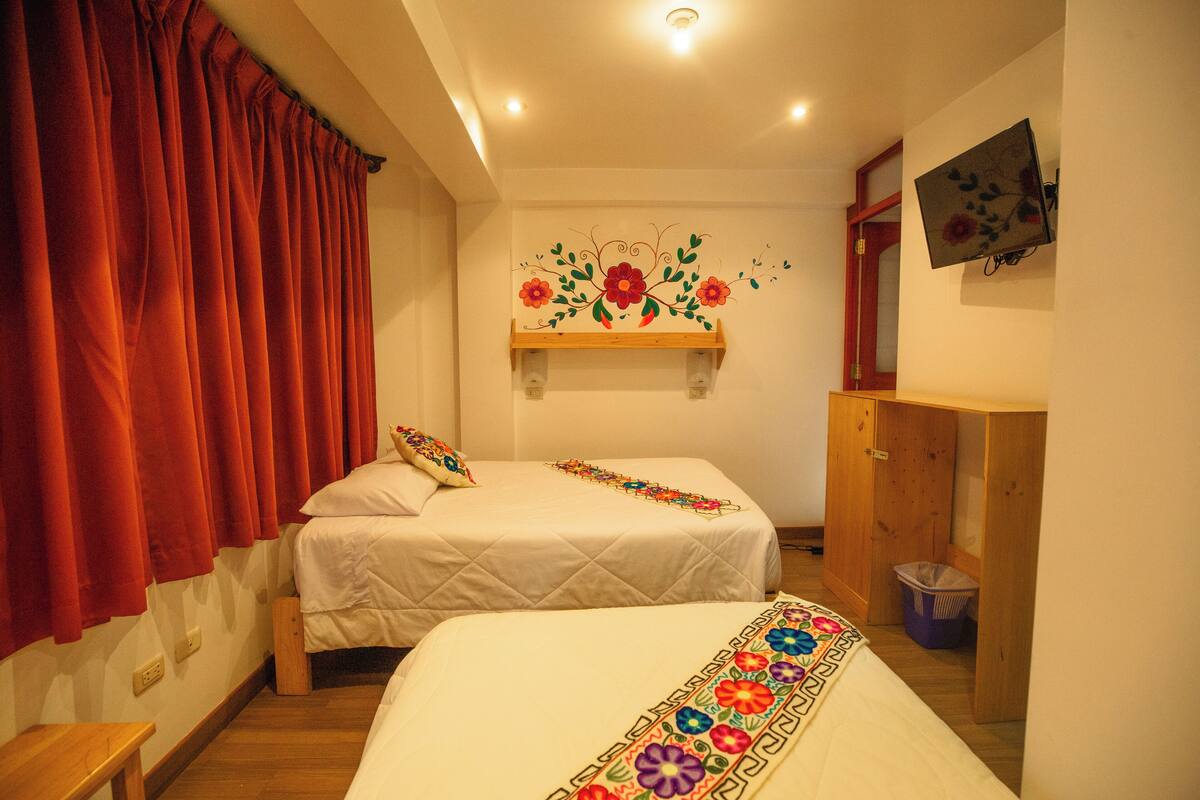 Un dormitorio con dos camas, cortinas rojas, una estantería de madera con adorno floral y un televisor en la pared.
