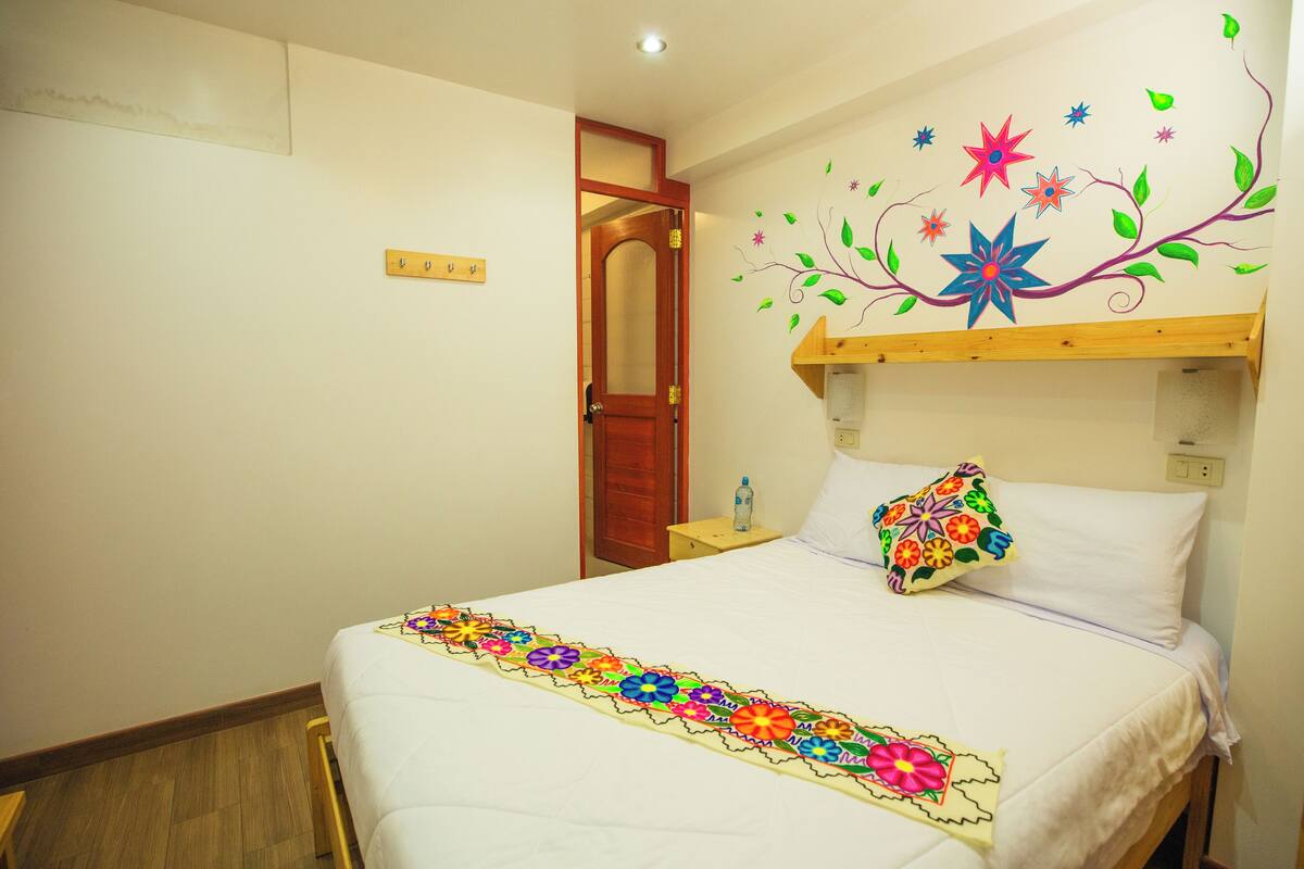 Una habitación de hotel con una cama, cabecero de madera, mesita de noche, botella de agua y una colcha de flores coloridas.
