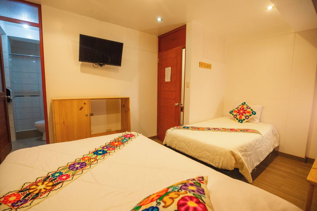 Habitación de hotel con dos camas, un armario de madera, televisor montado en la pared y baño a la vista a través de una puerta abierta.