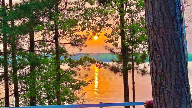 Vista para o lago com pôr do sol, árvores e área de estar ao ar livre.
