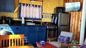 Cozinha compacta com armários azuis, fogão a gás, geladeira e área de refeições de madeira com mesa vermelha e duas cadeiras.