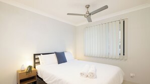Una cama bien tendida con ropa blanca y toallas enrolladas, una mesita de noche con un florero y una planta, un ventilador de techo y una ventana con persianas.