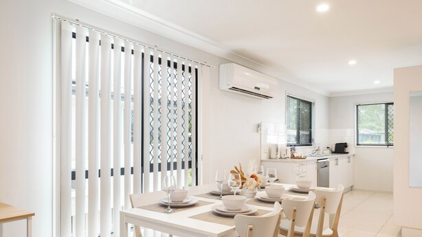 Una cocina moderna con armarios blancos, un comedor con mesa y sillas blancas, y vistas a través de una ventana con persianas.