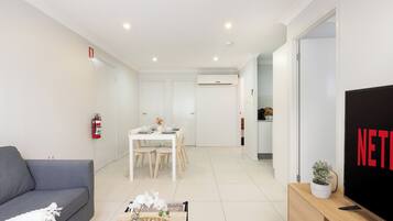 A modern living room with a grey sofa, a dining area with a white table and chairs, a TV on a wooden stand, and a fire extinguisher on the wall.