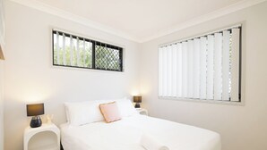 A bedroom with a bed, two bedside tables, a window with blinds, and a view of greenery outside.