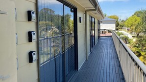A covered outdoor balcony with a wooden floor and glass doors, overlooking a residential area with trees and a clear sky.
