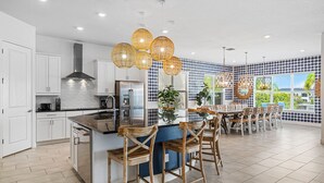 A modern kitchen with white cabinets, a central island with bar stools, and a dining area with a large window offering a view of greenery. - Legends Coastal Sanctuary: Private Pool, Theater, Near Disney (Kissimmee)
