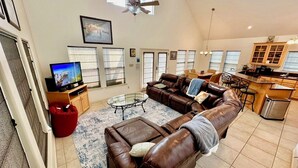 Een ruime woonkamer met een grote bruine leren bank, een glazen salontafel en een rode relaxfauteuil. Er is een televisie op een houten dressoir, een keuken met een bar en een eettafel met stoelen.
