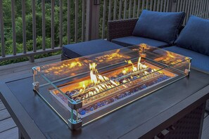 A glass-top fire feature on an outdoor table with a blue cushion and a wicker chair in the background.