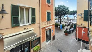 Una calle estrecha flanqueada por edificios, un pequeño espacio al aire libre con mesas y sillas, y una vista al mar al fondo.