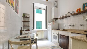 Una cocina compacta con azulejos blancos, una mesa de comedor con sillas, una ventana con contraventanas y un refrigerador pequeño.